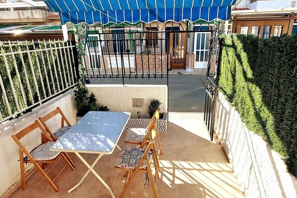 An outdoor patio with a table and chairs, a blue awning, a gated entrance, and a hedge.