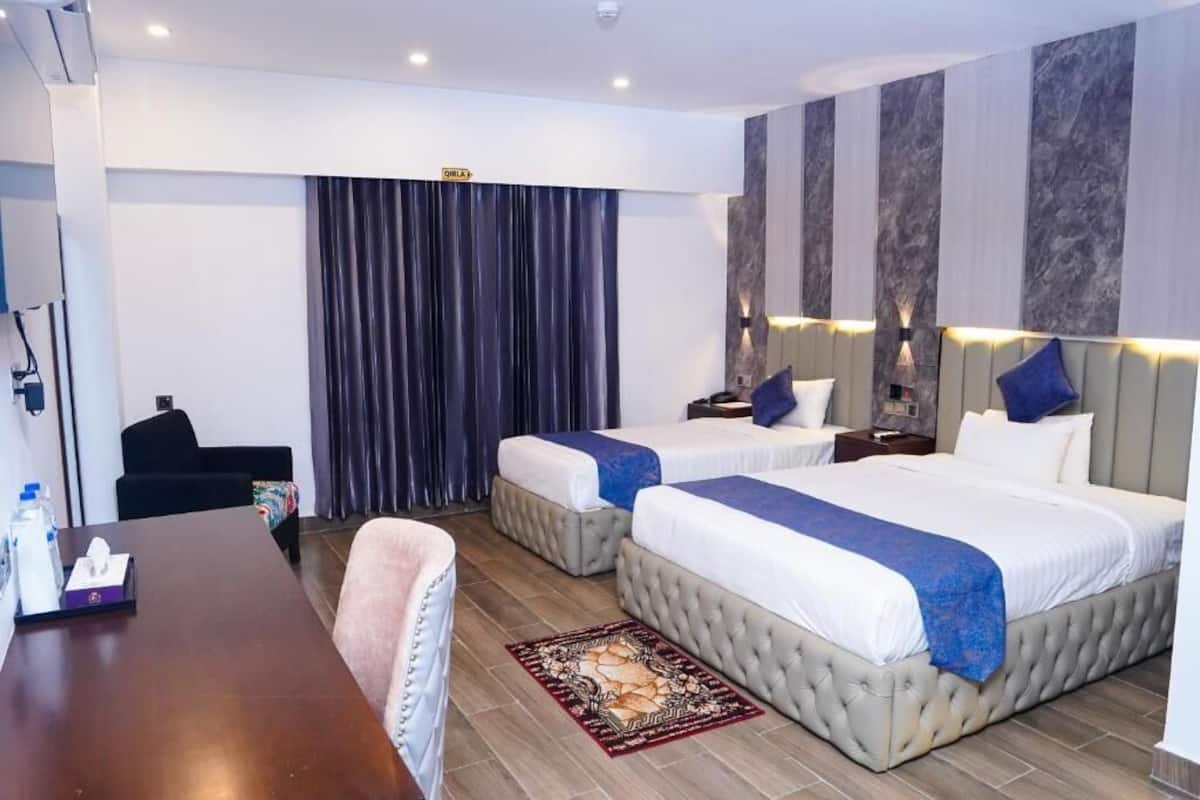 Une chambre d’hôtel avec deux lits, un bureau, une chaise, un rideau et un mur muni d’un luminaire.