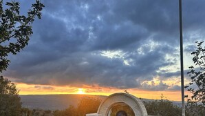 Une structure en pierre surmontée d’une icône religieuse, entourée d’une clôture en bois et de rochers, dominant un paysage où l’on aperçoit un coucher de soleil à l’horizon.