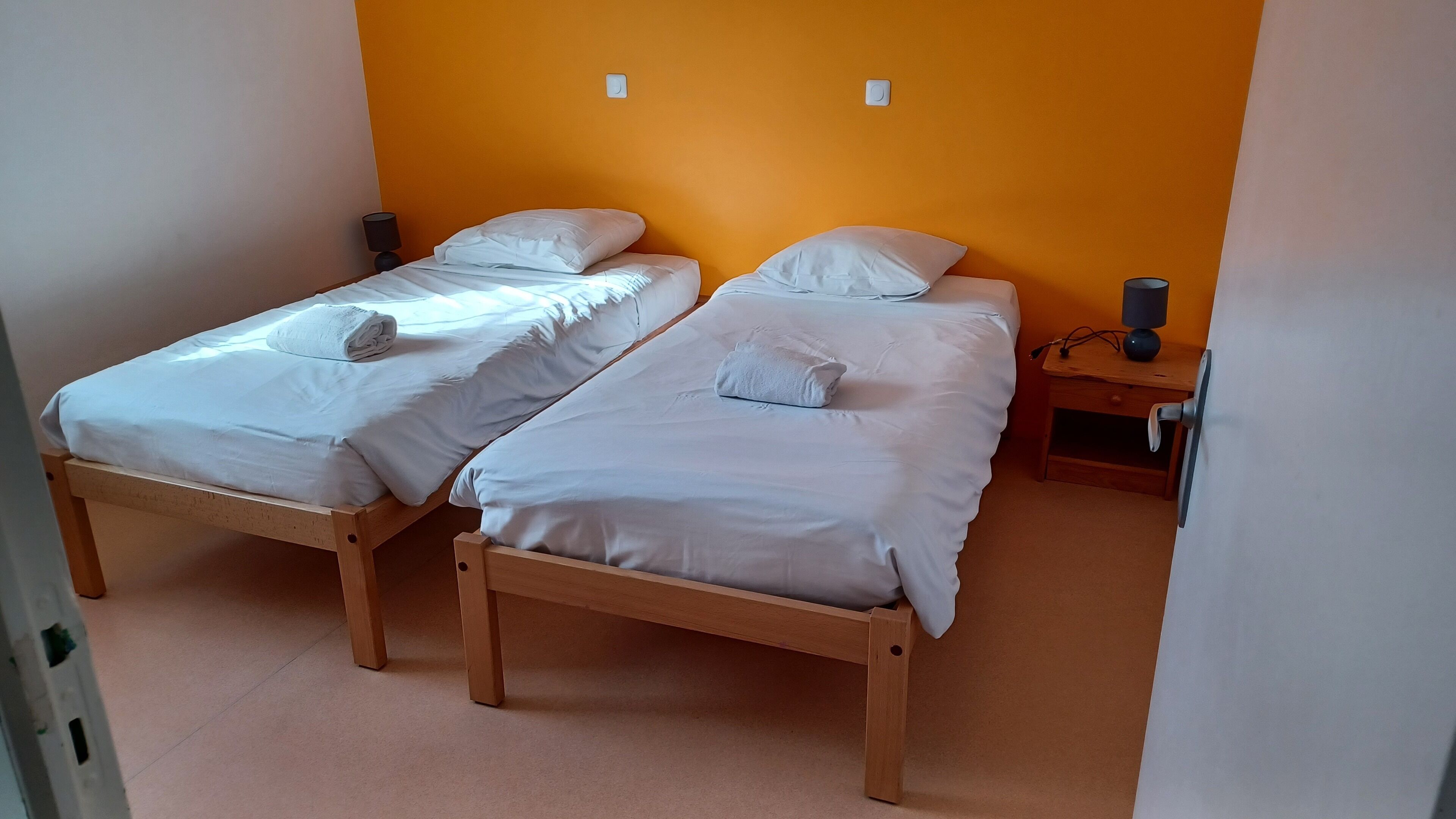 Dos llits individuals amb roba de llit i almoades blanques, una petita taula de nit de fusta amb una làmpada i una paret taronja sense adorns.
