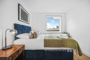 Quarto com cama, abajur, janela com vista para edifícios e quadro pendurado na parede.