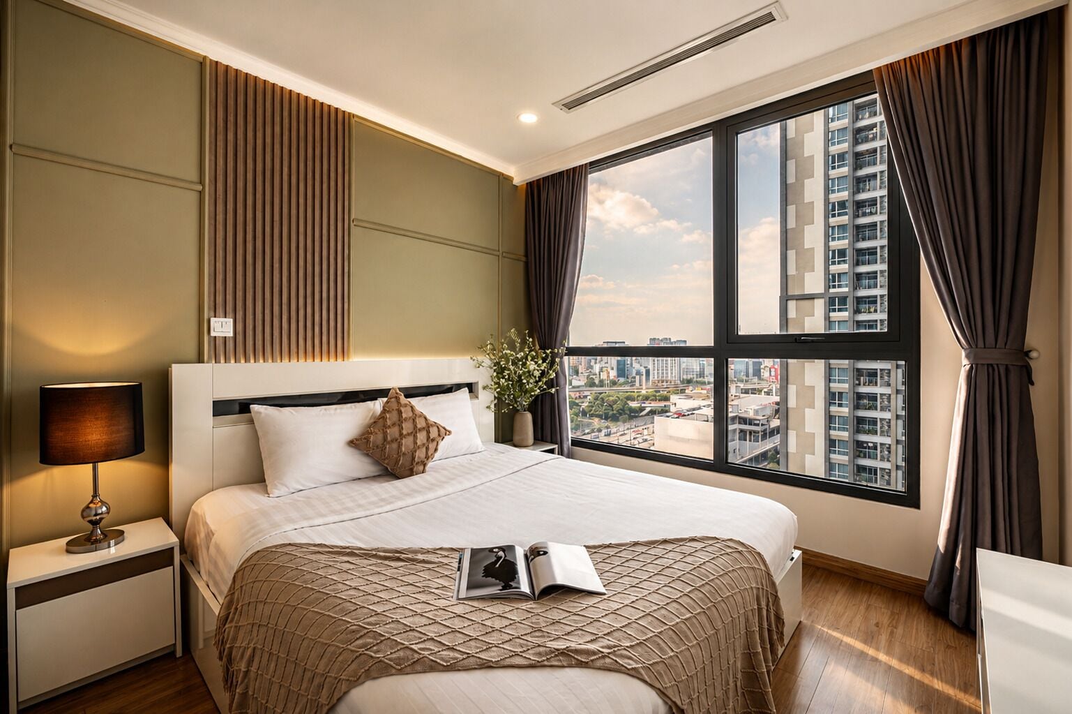 A hotel room with a large bed, a nightstand, a lamp, a window with curtains, and a city view.