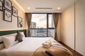 A hotel room with a large bed, a view of the city through a window, and framed artwork on the wall. - Vinhomes Skyline Apartment Saigon (Ho Chi Minh City)