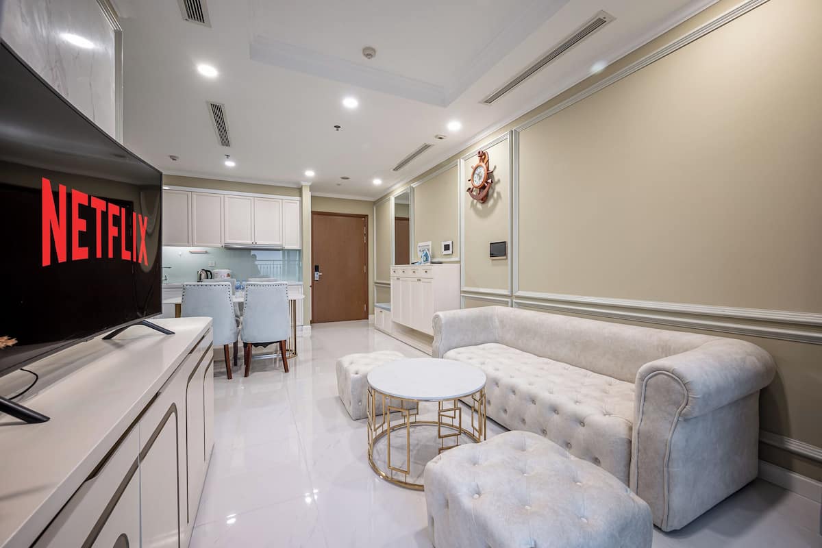 Una sala moderna con sofá, otomanes, una mesa de centro, televisor y una zona de cocina al fondo.