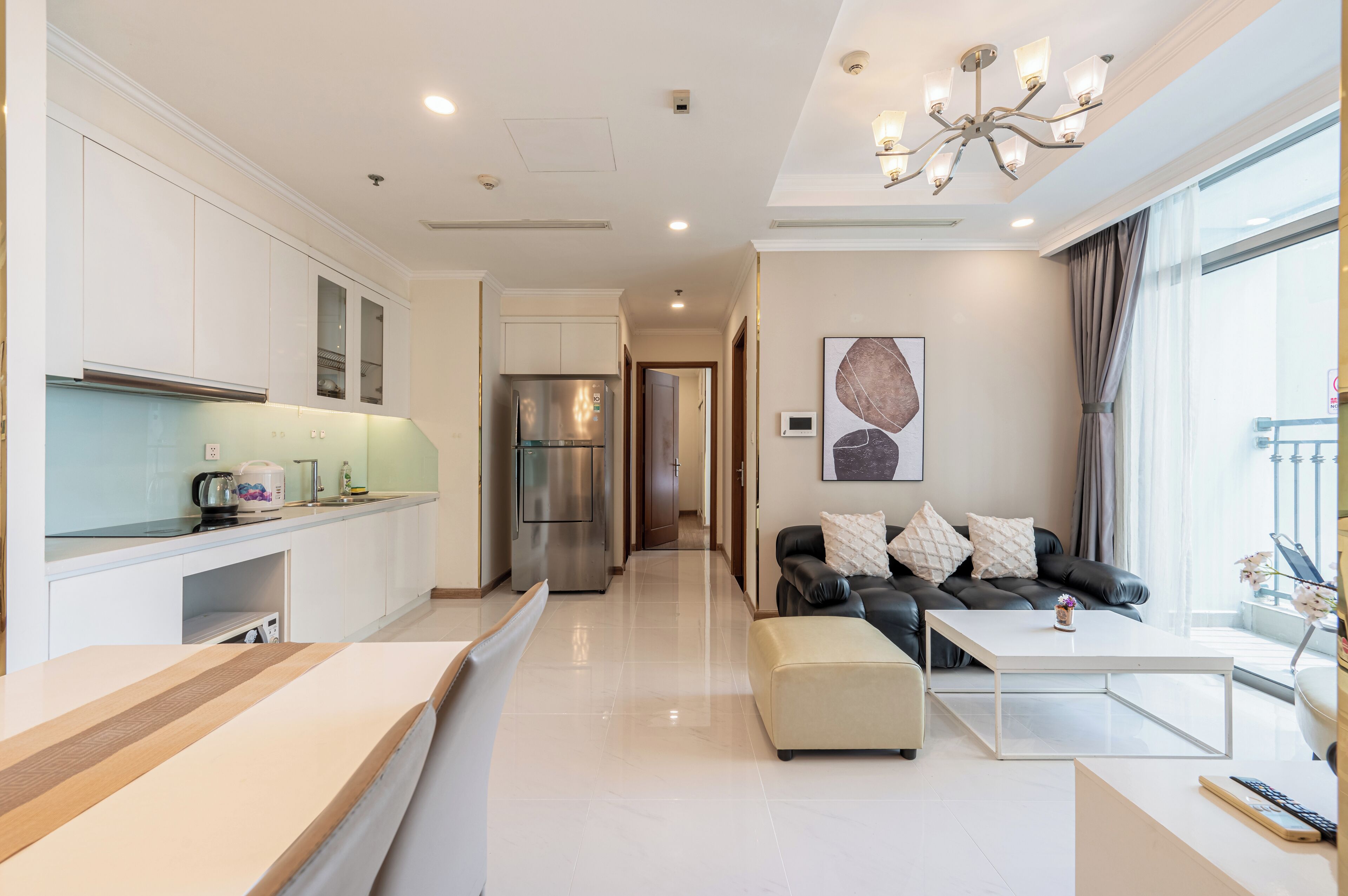 A modern living room with a sofa and ottoman, a kitchen with white cabinets and a refrigerator, and a large window with curtains.