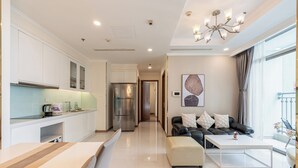 A modern living room with a sofa and ottoman, a kitchen with white cabinets and a refrigerator, and a large window with curtains. - Vinhomes Skyline Apartment Saigon (Ho Chi Minh City)