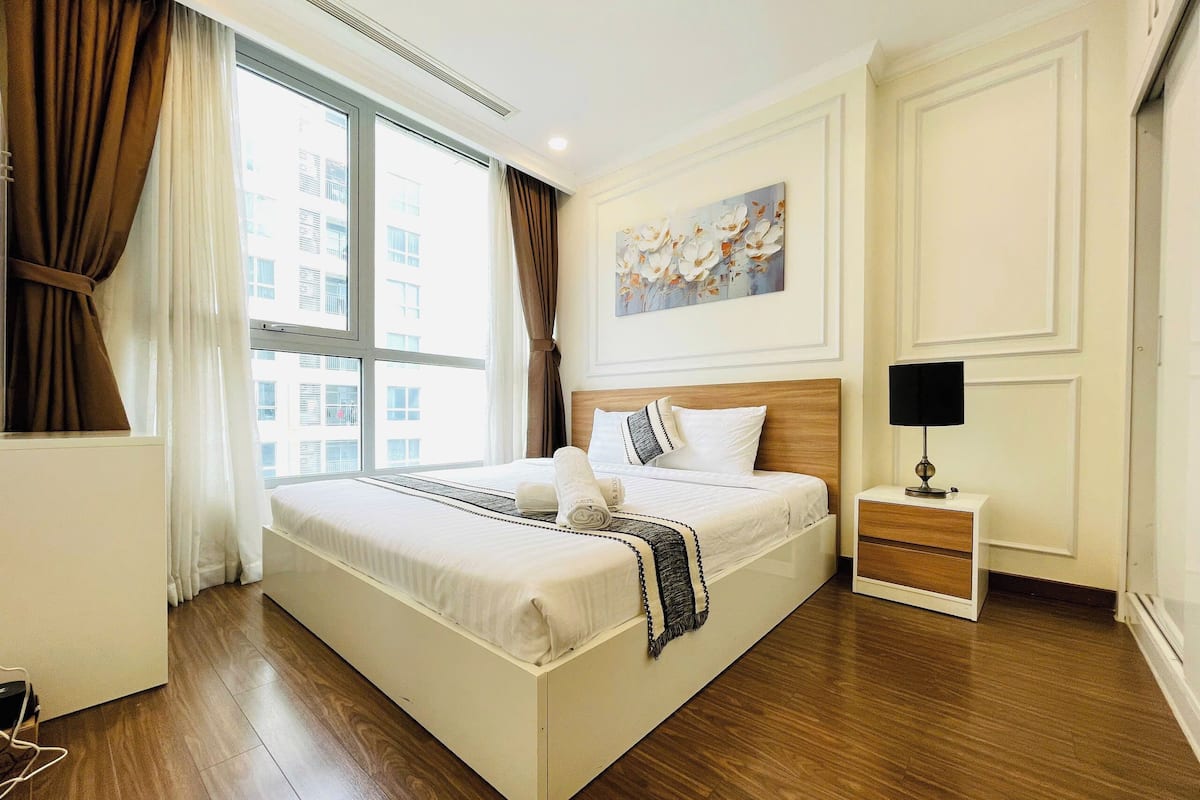 Habitación de hotel moderna con una cama grande, una mesita de noche con lámpara y vistas a la ciudad a través de amplios ventanales.