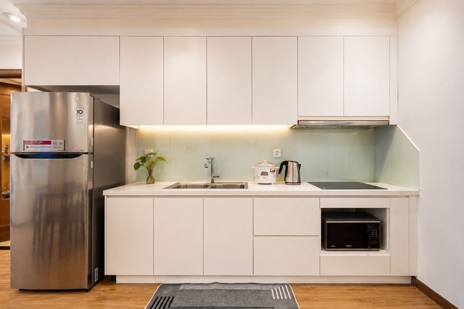 A modern kitchen with white cabinetry, a stainless steel refrigerator, a sink, a kettle, and an oven.