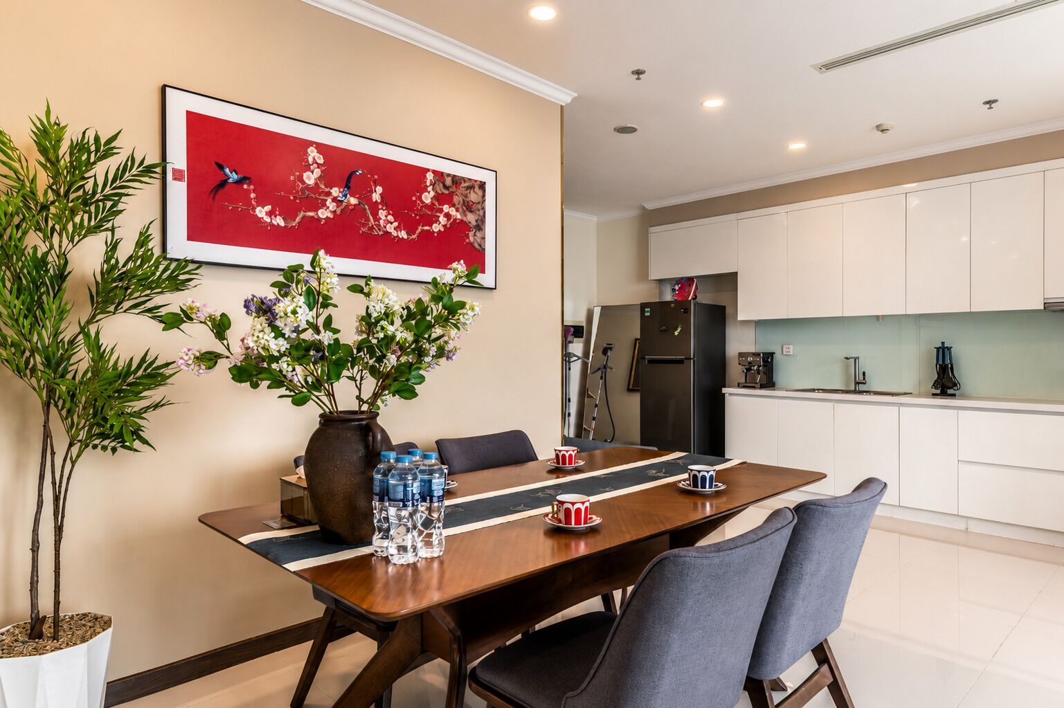 A dining area with a table, chairs, a vase with flowers, and a kitchen in the background.