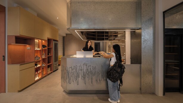 Un área de recepción de diseño moderno, con un mostrador de cristal y una persona interactuando con un miembro del personal. Hay estantes con varios artículos y una pared grande y texturizada sobre el mostrador.