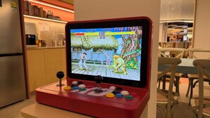 Una consola de videojuegos retro con una pantalla mostrando un juego, sobre una mesa de madera en una habitación con cocina al fondo.
