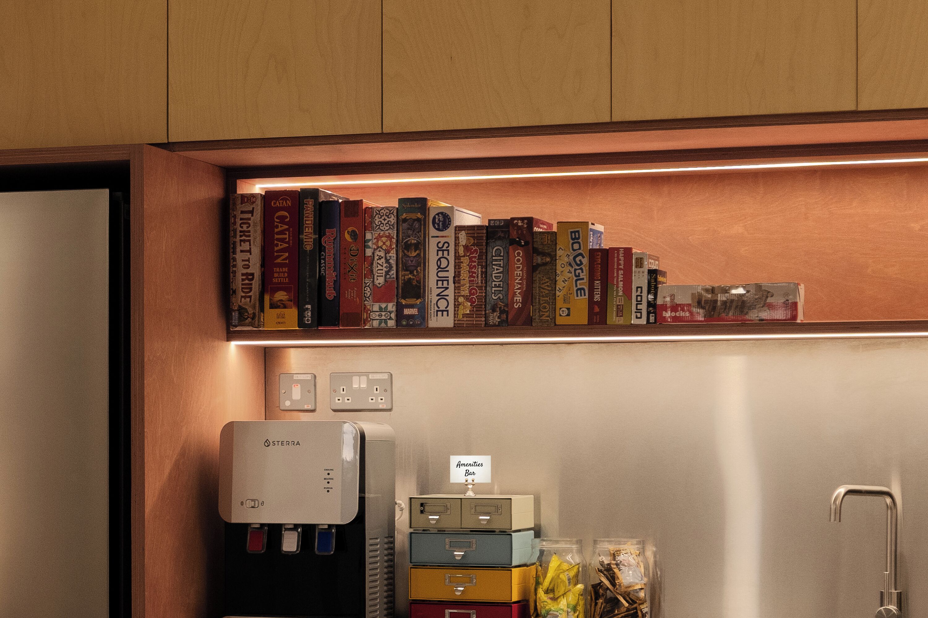 Una cocina con un refrigerador de acero inoxidable, un dispensador de agua y un estante con varios juegos de mesa.