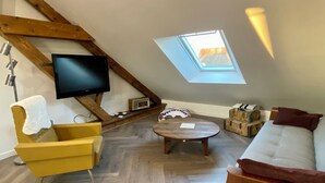 A loft-style room with a sloped ceiling, wooden beams, a television mounted on the wall, a sofa, a yellow armchair, a round wooden table, and a skylight. - Cosy loft in the heart of the Perche region (Val-au-Perche)