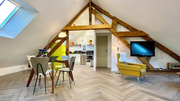 A spacious loft with a high ceiling, exposed wooden beams, a dining area with modern chairs, a kitchen with appliances, and a flat-screen TV mounted on the wall. - Cosy loft in the heart of the Perche region (Val-au-Perche)