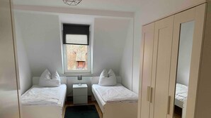 A small, well-lit room with two single beds, a desk, a chair, a radiator, and a window with a view of a building outside. - Stay in a Historic Half-Timbered House (Nürnberg)