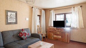 Ein Wohnzimmer mit einer Couch, einem Fernseher, einem Schrank und einem Fenster mit Vorhängen.