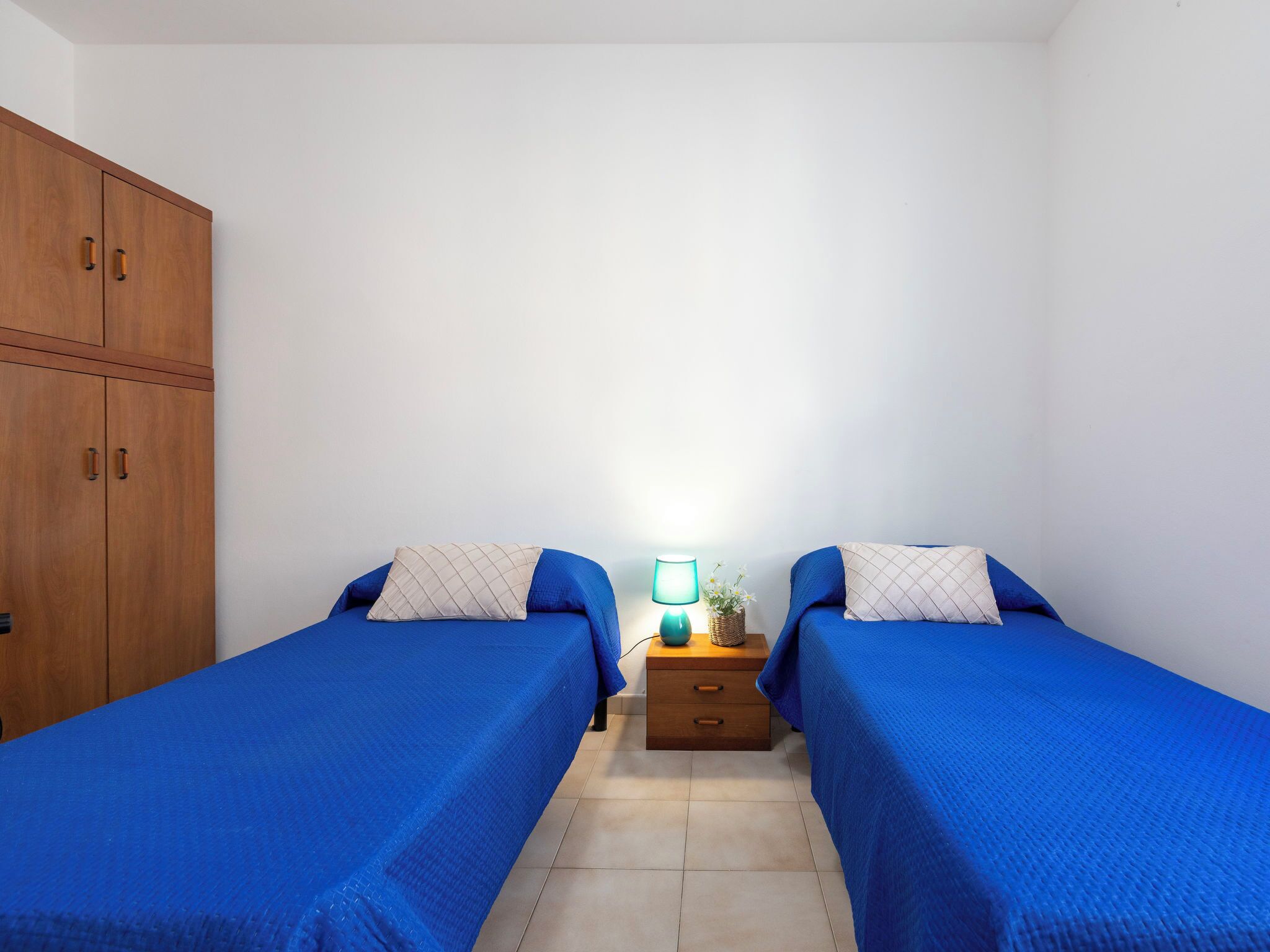 A bedroom with two beds, a wooden wardrobe, a nightstand with a lamp, and a small plant.