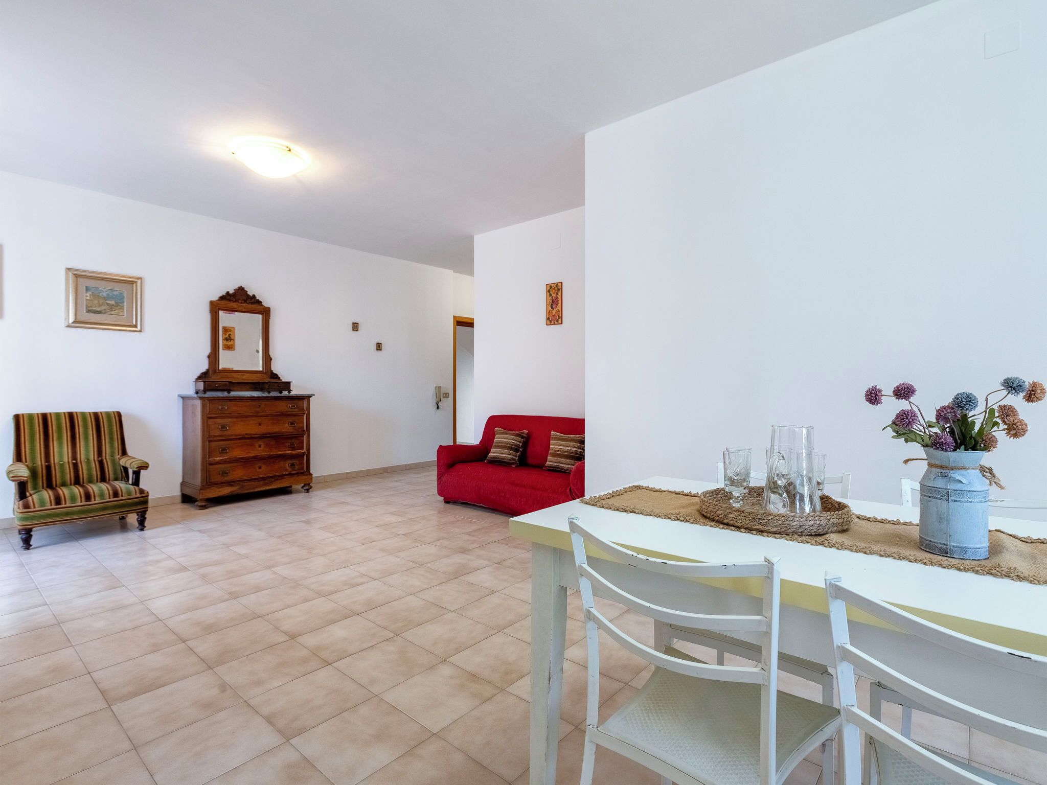 A spacious living area with a tiled floor, a wooden dresser, a striped armchair, a red sofa, and a dining table with chairs.