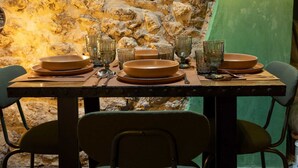 Uma mesa posta para refeição, com pratos, copos e talheres, ao fundo de uma parede de pedra.