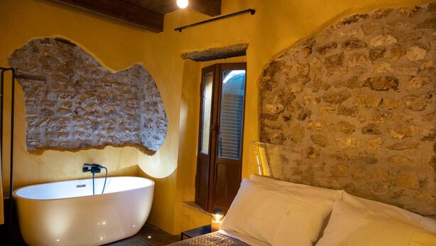 Quarto com uma banheira grande, cama com roupa de cama branca e paredes de pedra.