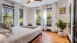 A bedroom with a bed, a ceiling fan, large windows with curtains, a potted plant, and a painting on the wall. - Sanctuary on 3rd | Walk to UF • Parking • Grill (Gainesville)
