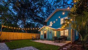 A house with a blue exterior, a wooden fence, a patio with chairs, and string lights. - Sanctuary on 3rd | Walk to UF • Parking • Grill (Gainesville)