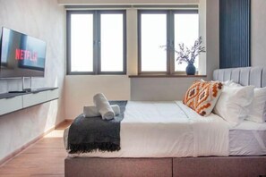 A modern hotel room with a bed, a TV displaying 'NETFLIX,' a window with a view, and a vase with branches. - Riveria City near KL Sentral with Sky Infinity Pool by COBNB (Kuala Lumpur)
