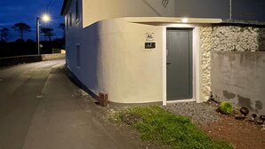 Un edificio de dos pisos con fachada blanca y puerta gris, un pequeño letrero que indica una entrada accesible y un alumbrado público al fondo.