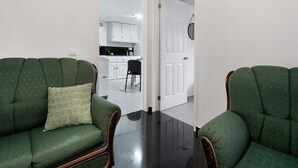 Una habitación con dos sillones tapizados de verde, un cojín estampado y una cocina visible a través de una puerta abierta.