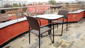 A balcony with a table and two wicker chairs, overlooking a cityscape with buildings and trees.