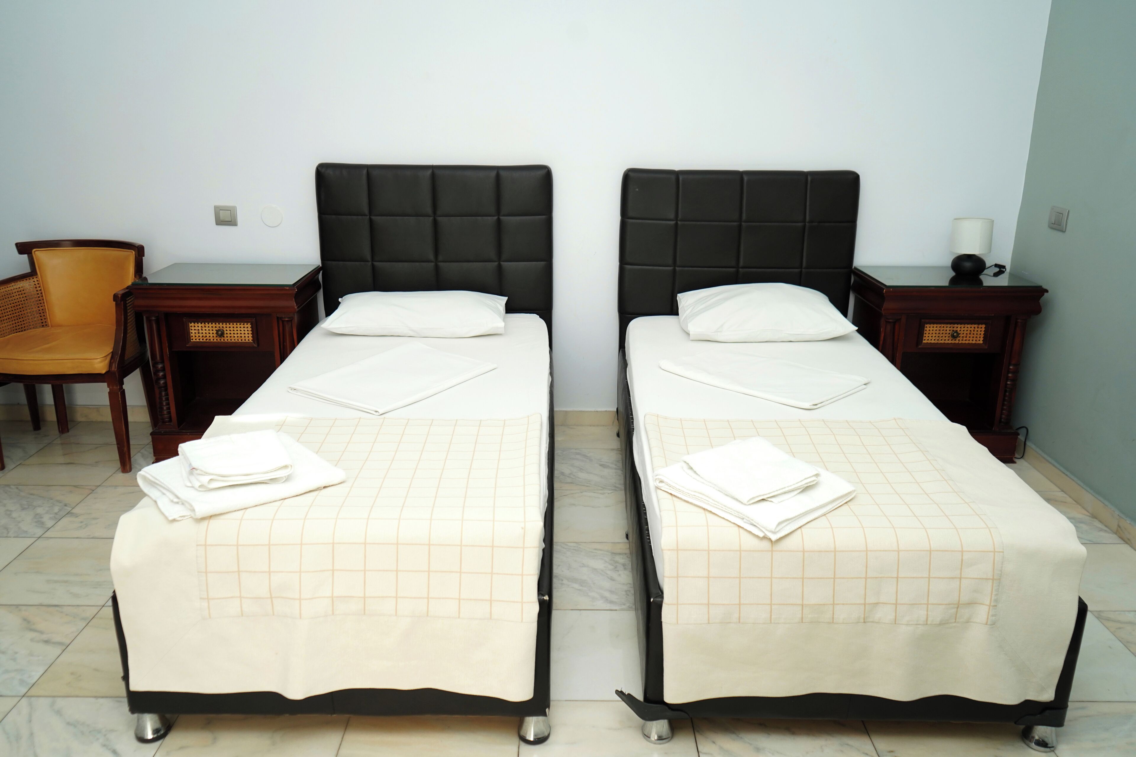 Two single beds with black headboards, white linens, and a checkered bedspread. There are wooden nightstands with lamps and a wicker chair.