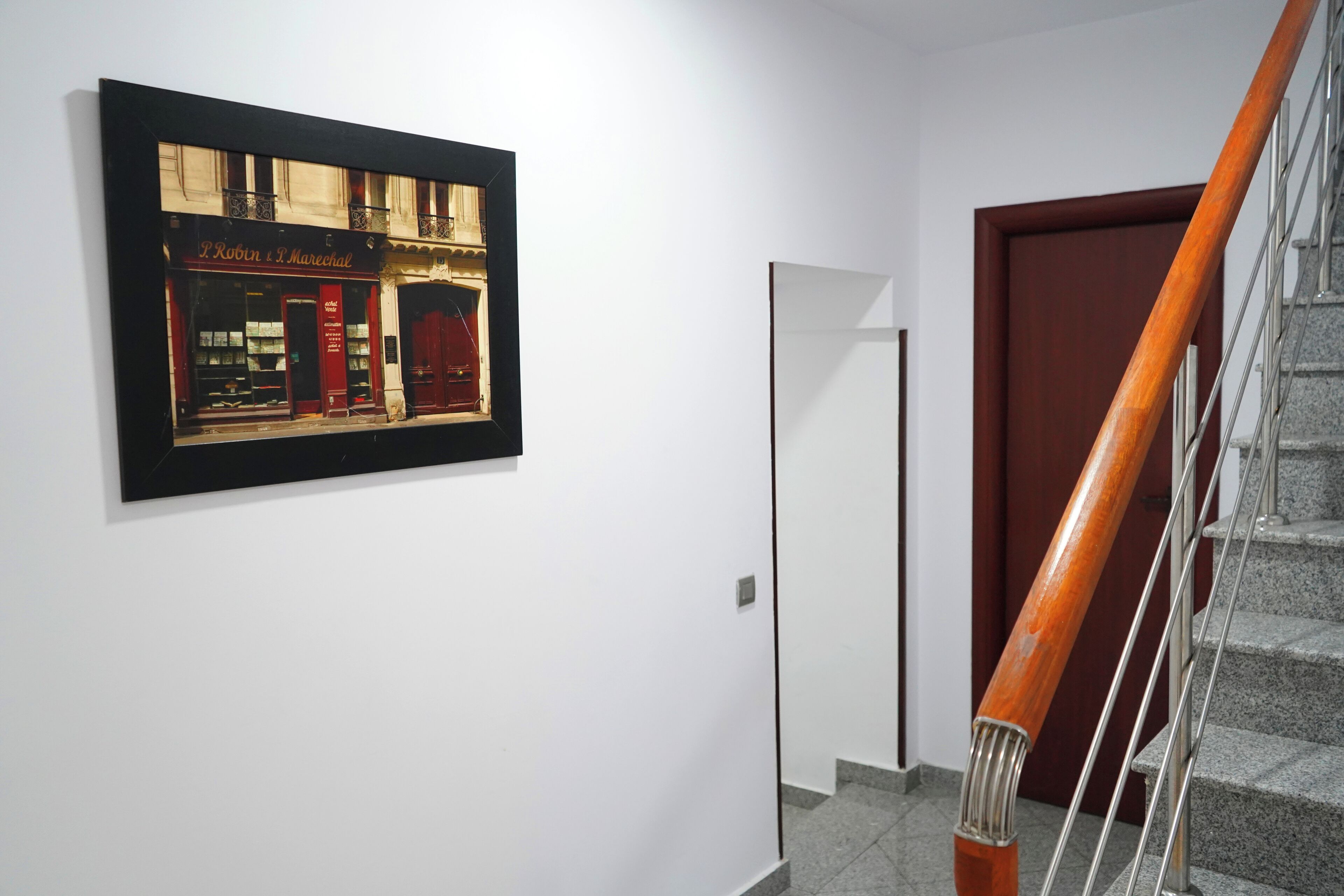 A hallway with a framed picture of a storefront, a wooden door, and a staircase with a wooden handrail.