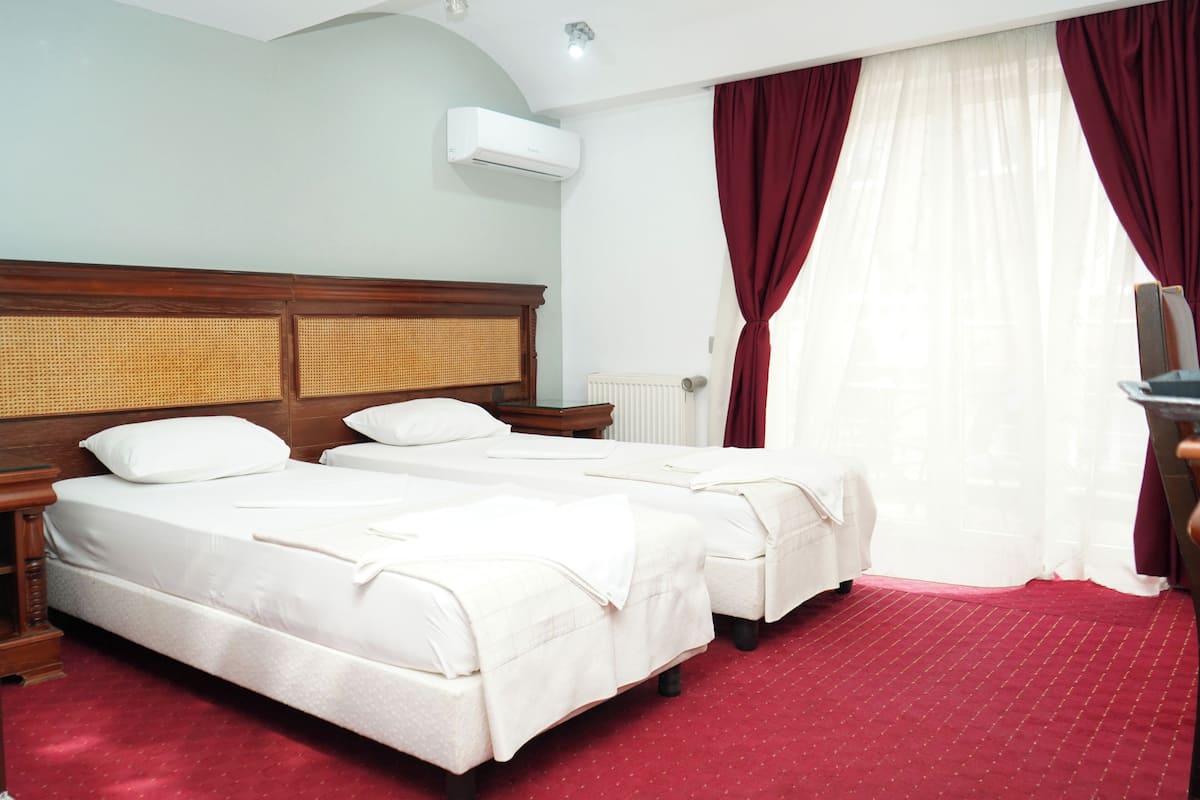 Habitación de hotel con dos camas, cabecera de madera, radiador y ventana con cortinas rojas.