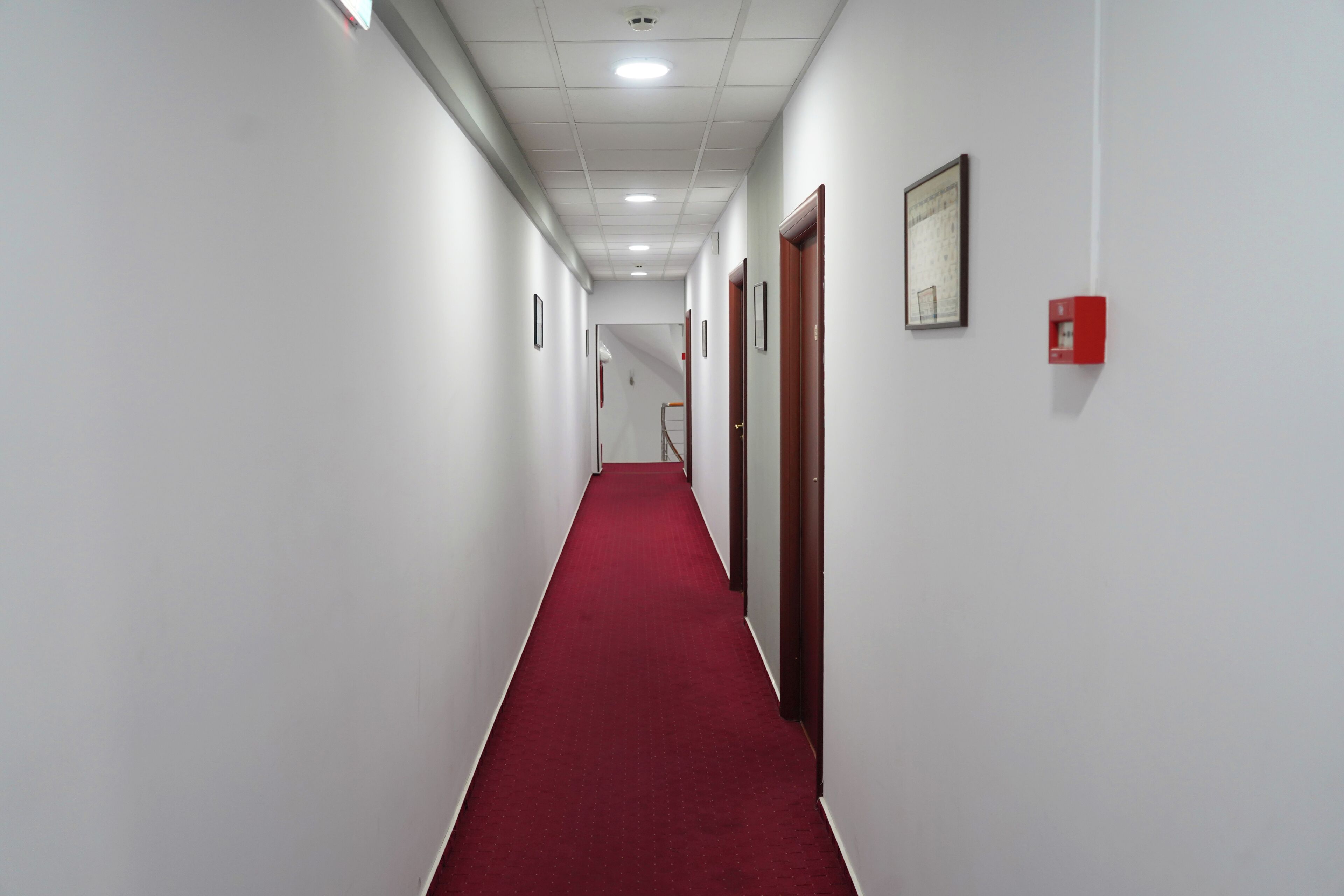 A long, narrow hallway with white walls and a red carpet, leading to a door at the end.