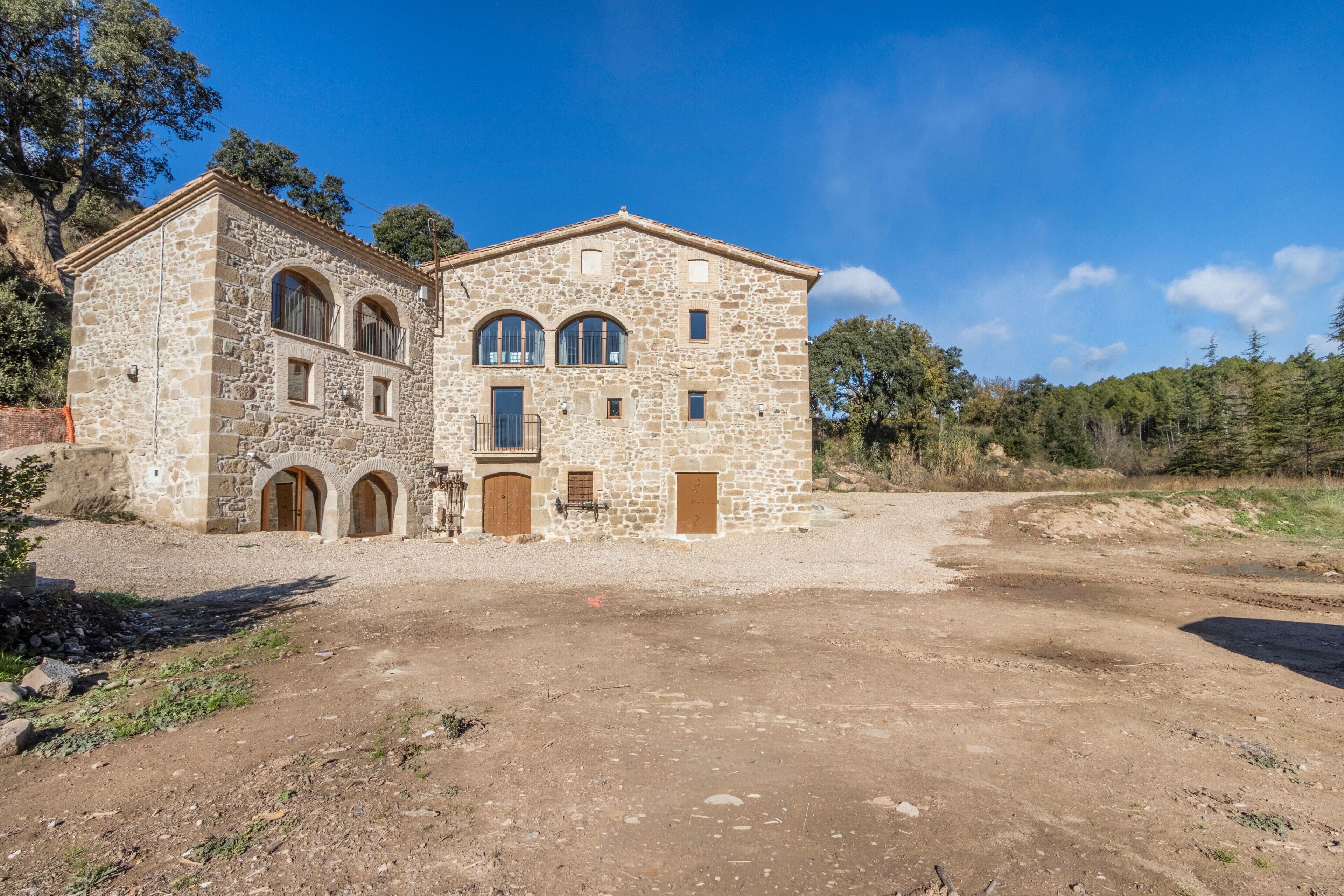 A stone building with arched windows and doors, surrounded by a dirt area and trees.