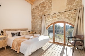 Un dormitorio con pared de piedra, una cama grande, una mesita de noche, una silla y vistas a través de un ventanal.