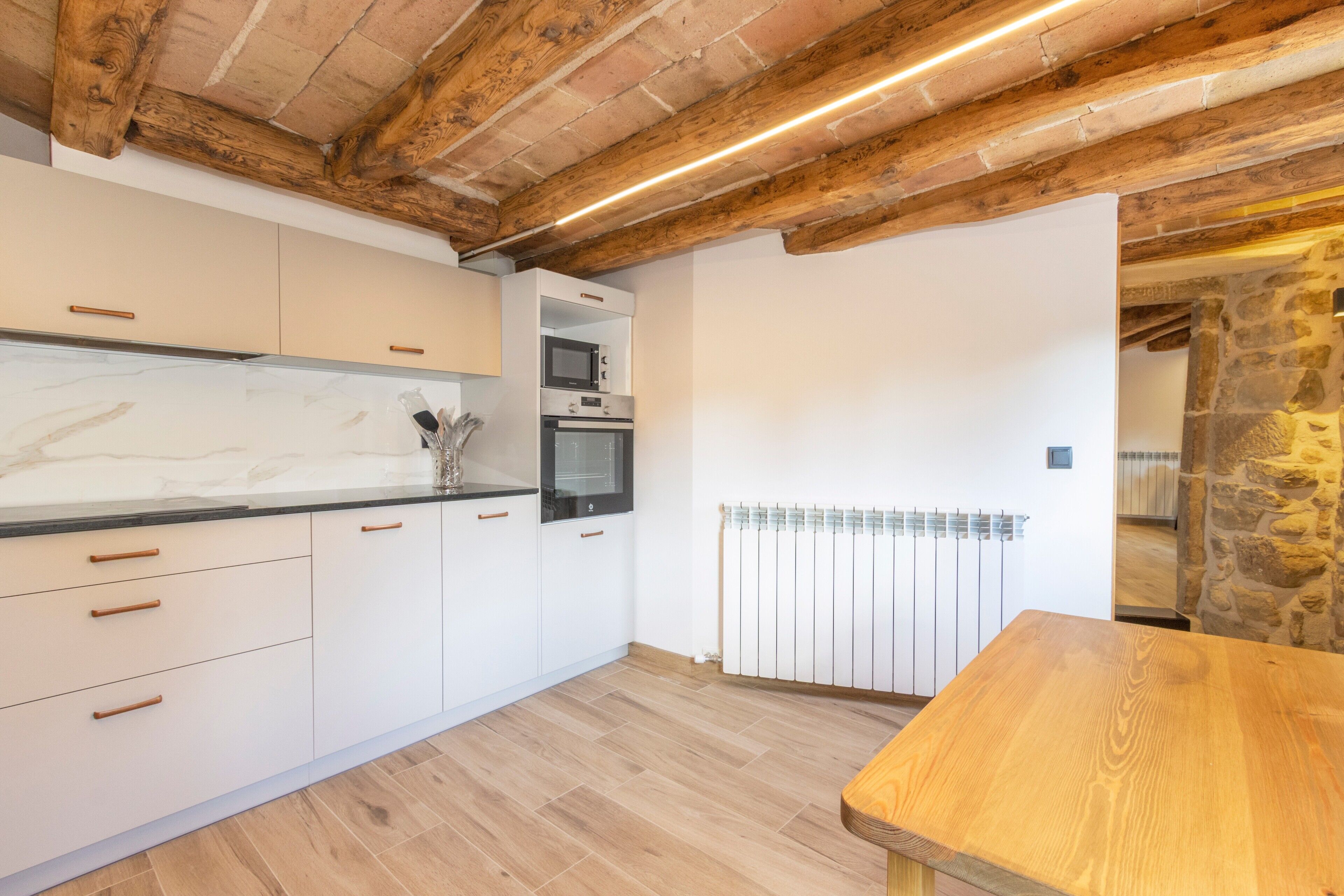A modern kitchen with white cabinets, wooden ceiling beams, and a stone wall.