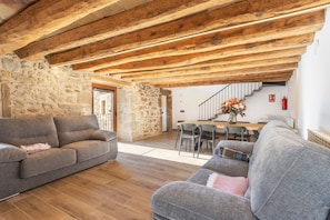 Amplia sala de estar con pared de piedra, vigas de madera, un sofá y un comedor con sillas y una mesa.