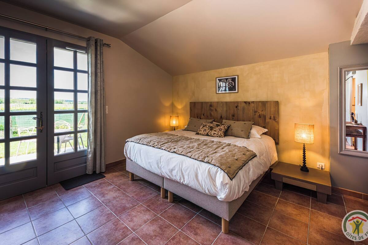 Ein Schlafzimmer mit einem großen Bett, zwei Nachttischlampen, einem Holz-Kopfteil und Blick auf einen Garten durch eine Schiebetür.