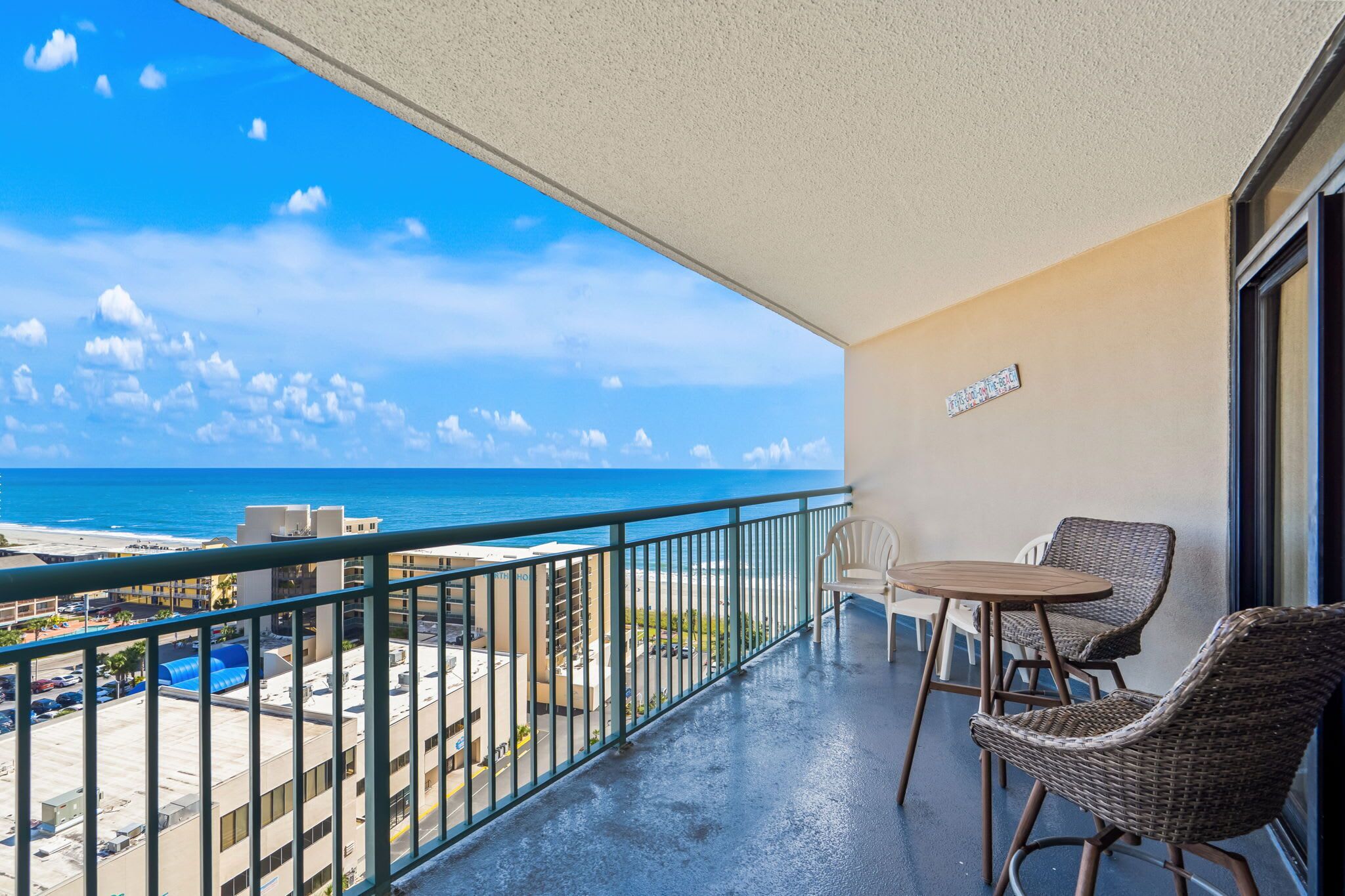 A balcony with wicker chairs and a table, overlooking a beach and ocean.