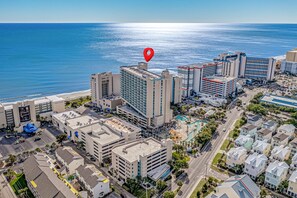 Ett flygbildsbild av ett hotellområde vid kusten med flera byggnader, inklusive ett framstående hotell med en pool på taket och utsikt över havet.