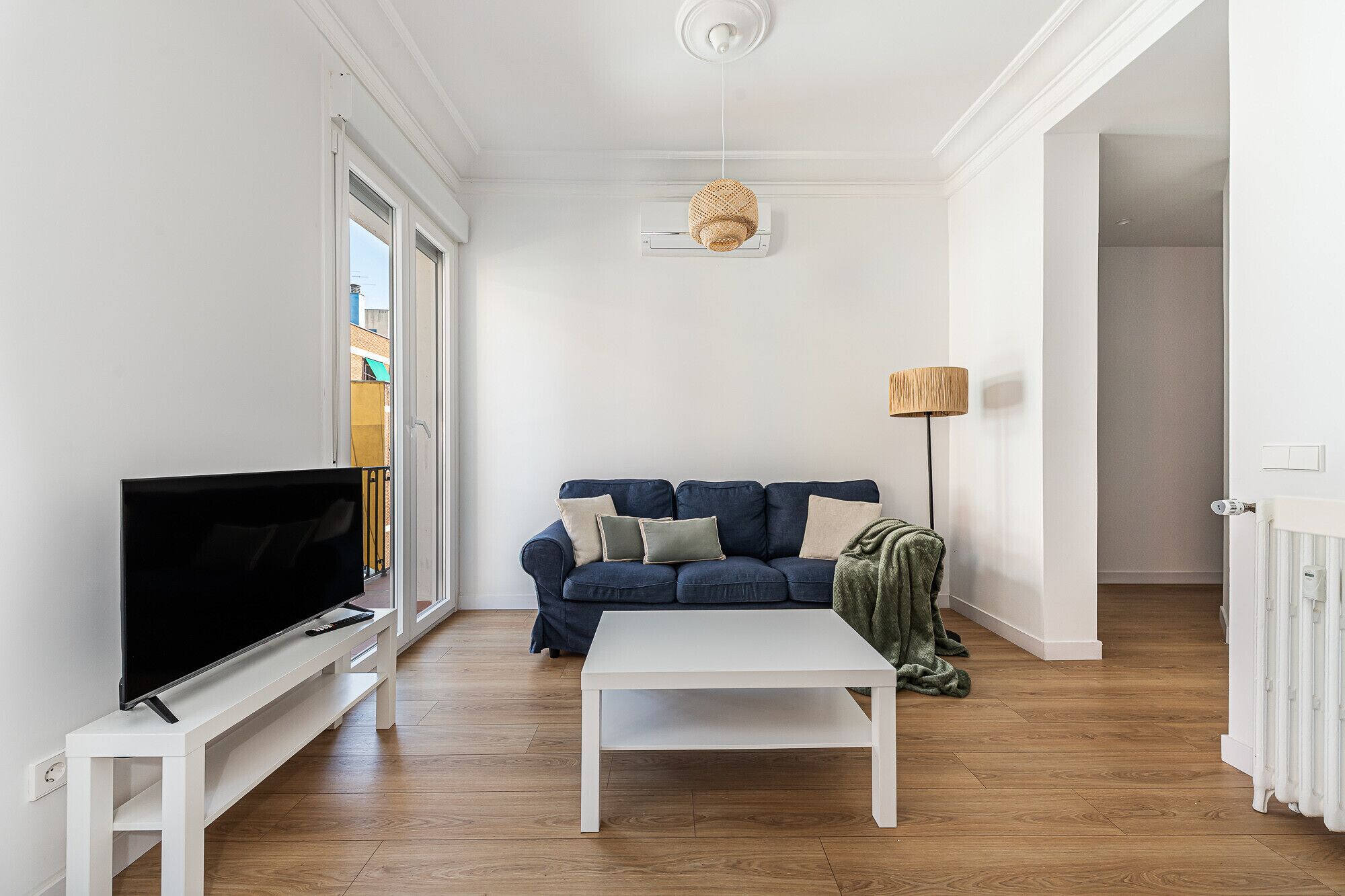 Een woonkamer met een televisie op een witte zuil, een blauwe bank met kussens, een witte salontafel en een lamp.