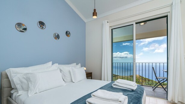 A bedroom with a bed, a blue blanket, and a view of the sea through a balcony. - Madeiran Vernacular Villa • Sunset Views • Garden • Heating (Ponta do Pargo)