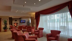A spacious living area with red upholstered sofas, a fireplace, a flat-screen TV, and a large window with sheer curtains. - Albergo Conte Max (Capracotta)