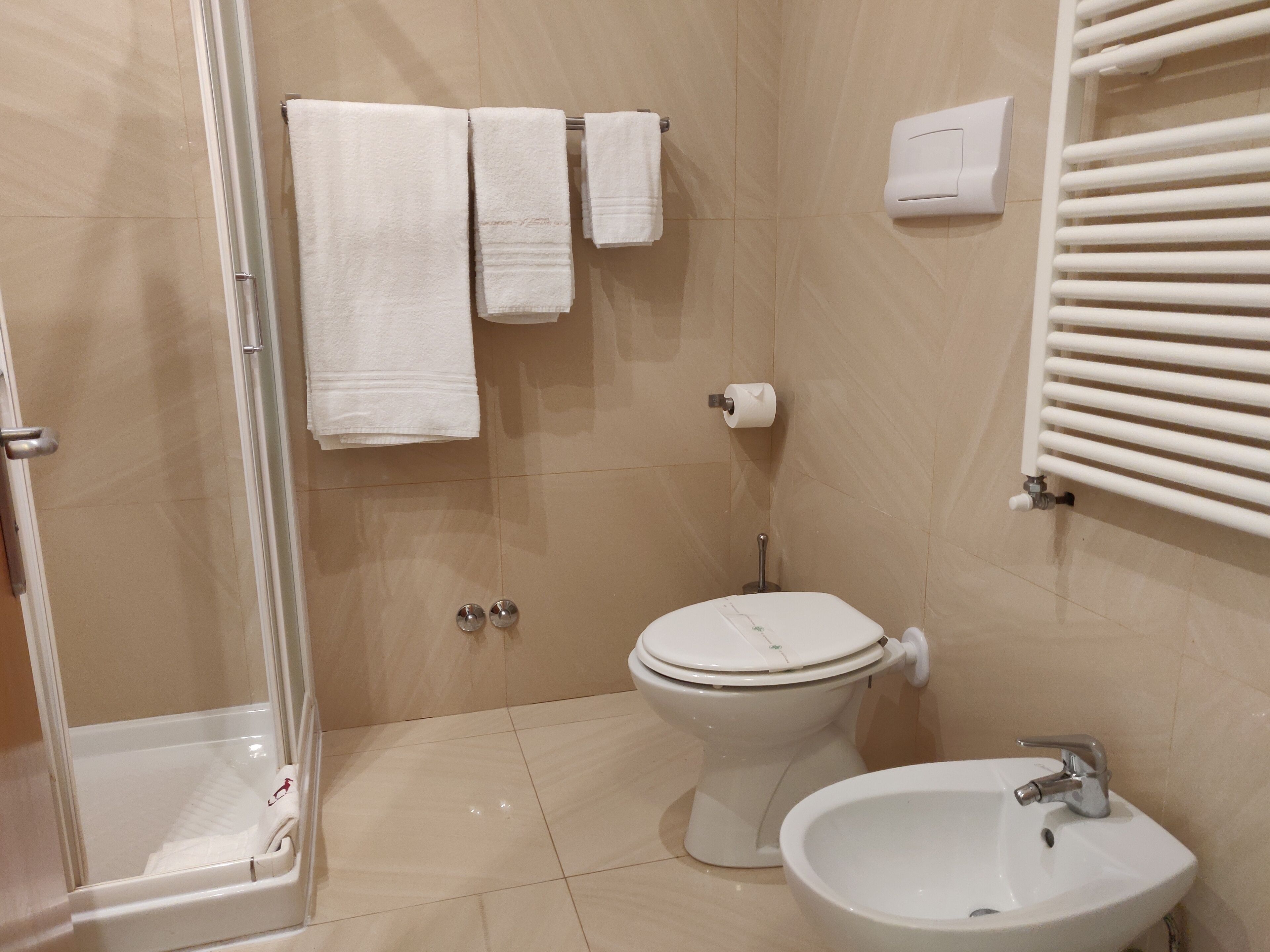 Ein Badezimmer mit Toilette, Waschbecken und Dusche. Handtücher hängen an einem Ablagegestell und eine Toilettenpapierhalterung ist an der Wand angebracht.