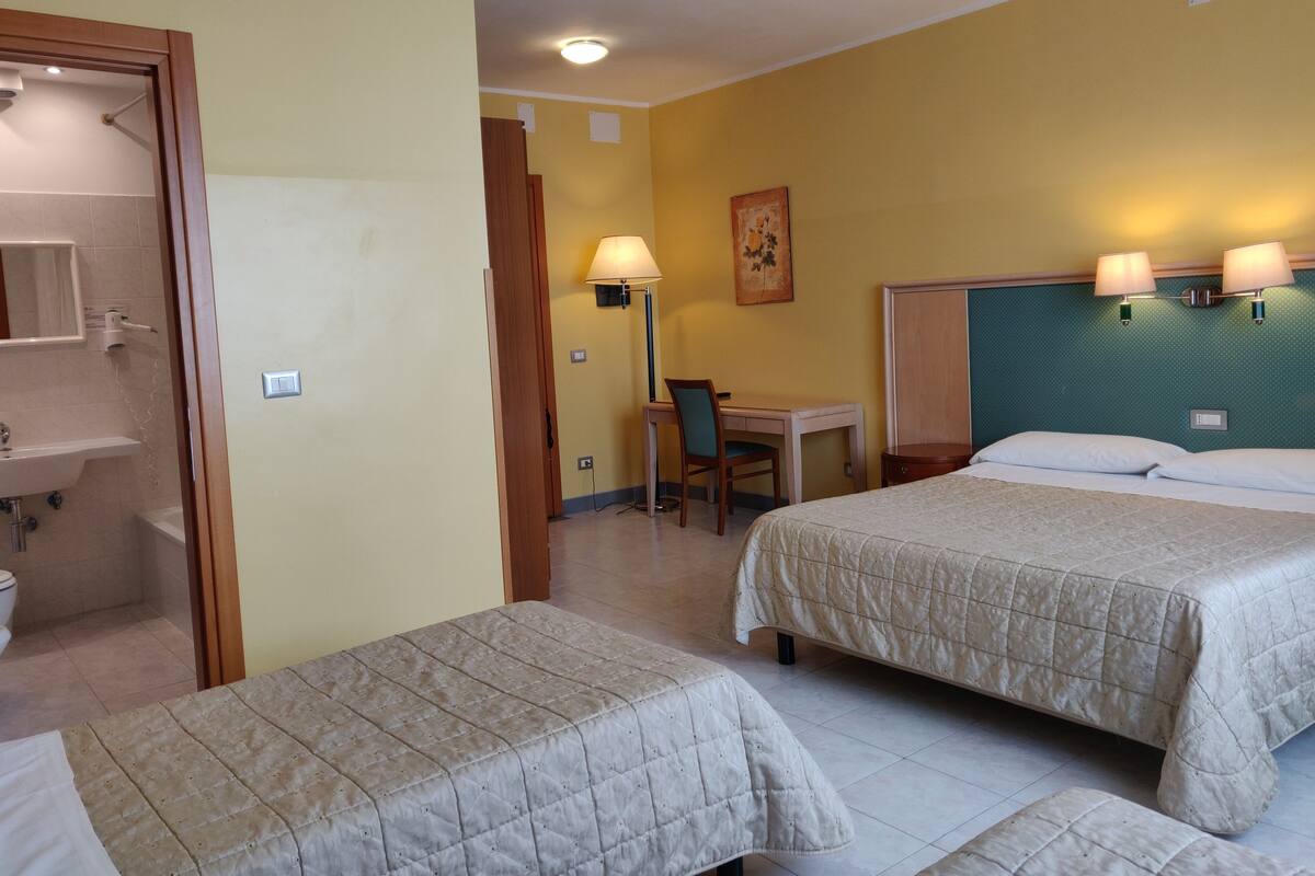 Hotellihuone, jossa on kaksi sänkyä, työpöytä, tuoli, lamppu, taulu sekä kylpyhuone, jossa on pesuallas ja wc.