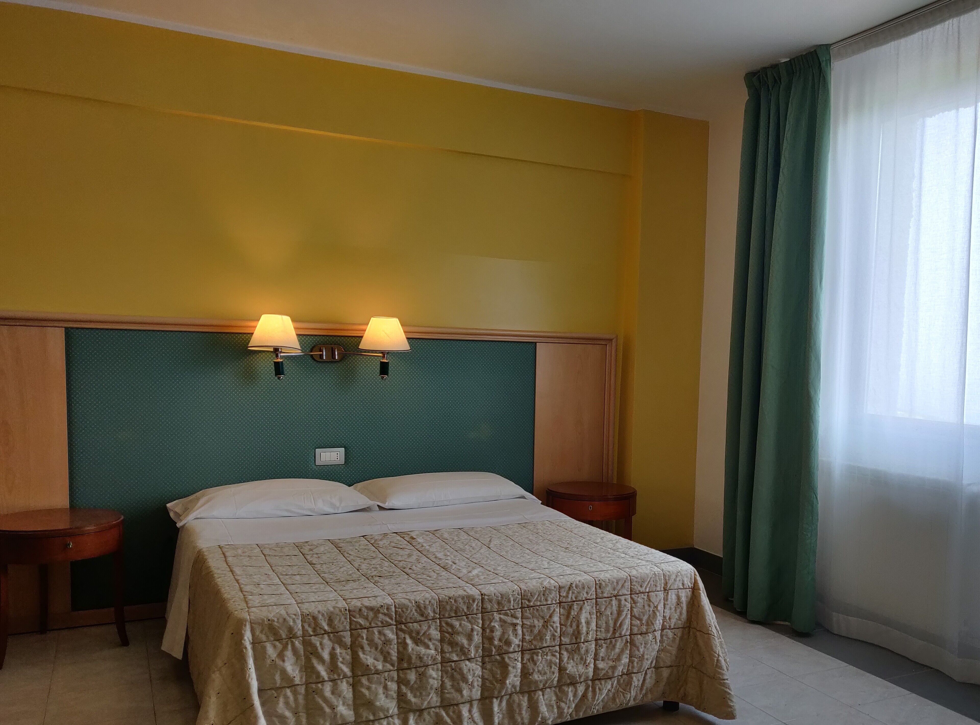Ein Hotelzimmer mit einem Bett, zwei Nachttischlampen, einem Nachttisch und einem Fenster mit Vorhängen.