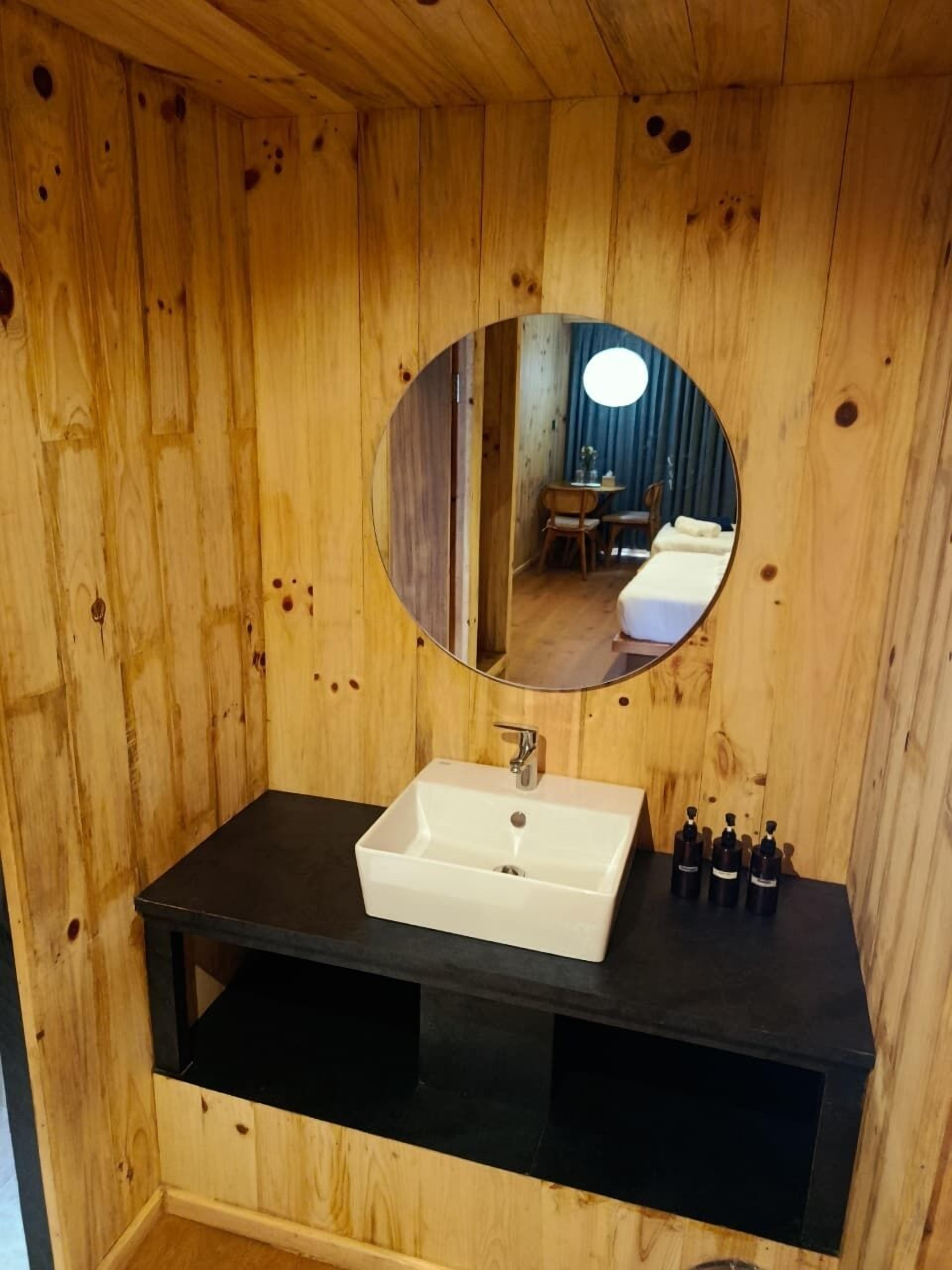 Une salle de bain en bois avec un lavabo blanc, un plan de toilette noir et un miroir rond.