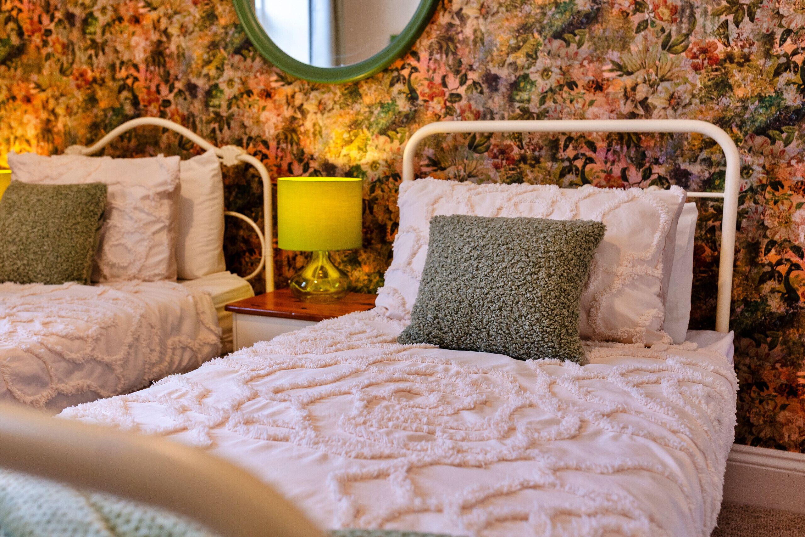 A bedroom with two beds, floral wallpaper, a round mirror, and a bedside lamp.