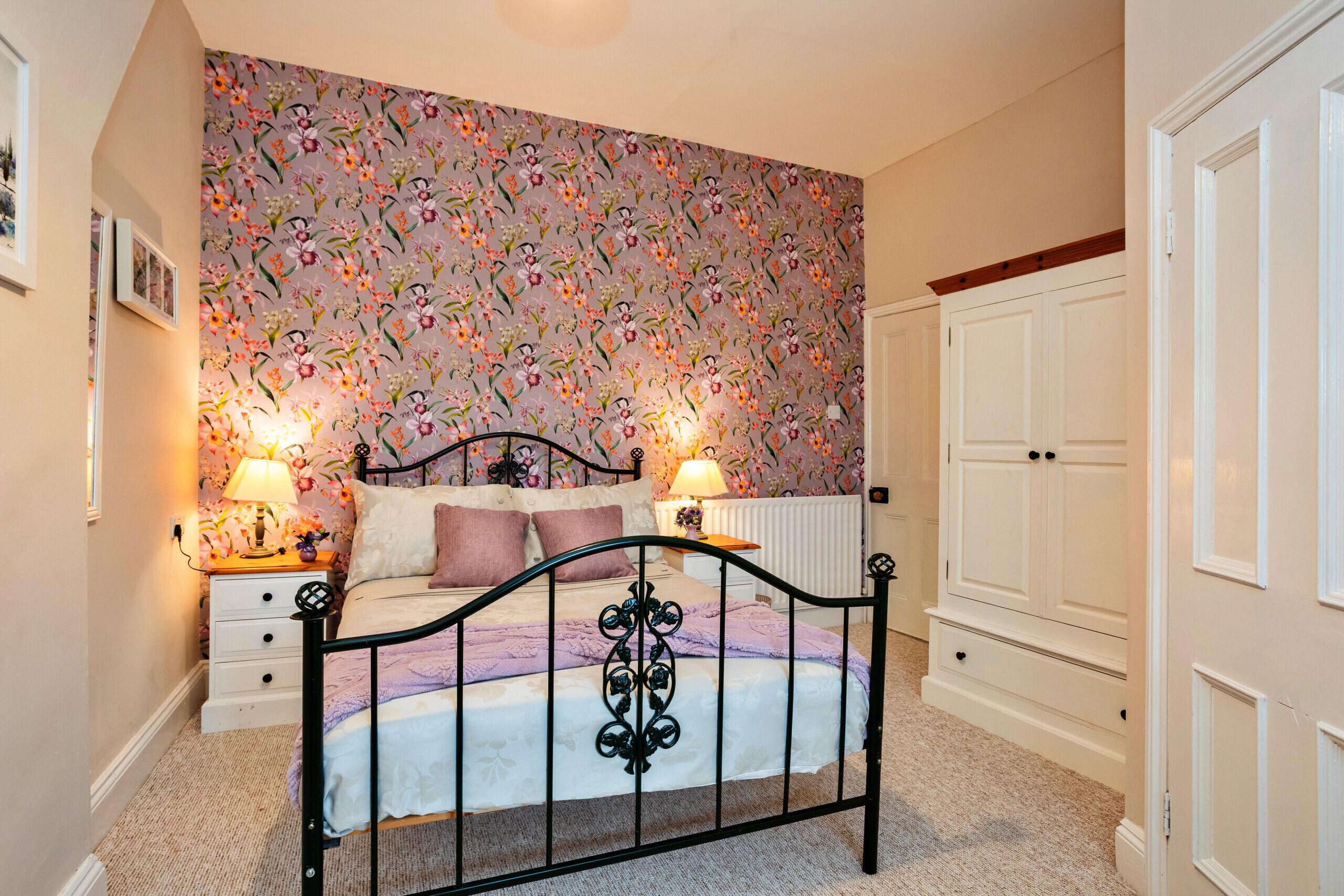 A bedroom with a floral wallpaper, a bed with a black metal frame, two bedside tables with lamps, and a white wardrobe.